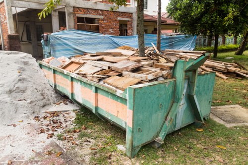 A large green metal skip positioned on a grassy area beside a construction site, filled with irregularly stacked wooden planks and pallets that vary in size and thickness, with some showing signs of weathering and rough finishes. The skip's sides are reinforced with orange banding, and its front features a partially open lifting mechanism. In the background, there is a partially constructed brick building with exposed brickwork and a blue tarpaulin covering part of the scaffolding, along with a wooden utility pole supporting overhead wires. To the right, mature trees with green foliage provide shade and a natural backdrop, while a paved driveway or footpath runs nearby, indicating a residential or semi-urban setting. The environment is well-lit with natural daylight, emphasizing the textures and colours of the discarded timber and construction debris, which is typical of rubbish clearance work conducted by specialists like Twickenham House Clearance in local areas such as Twickenham or surrounding TW postcodes.