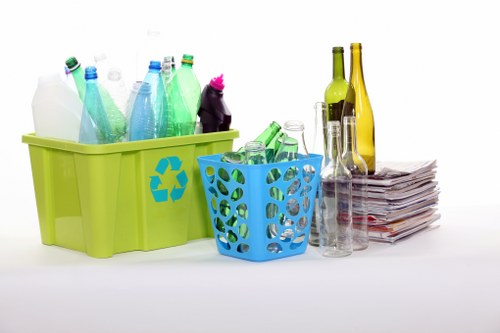 A collection of recyclable waste bottles and glass containers on a white surface, including a lime-green plastic bin with a blue recycling symbol filled with various plastic bottles in shades of white, green, and blue, alongside a smaller blue plastic basket containing additional plastic bottles and containers. To the right, there are empty glass bottles in yellow and green hues, and a small pile of flattened newspapers or magazines. The scene appears to be a workplace or home environment where rubbish removal services by Twickenham House Clearance could assist in the collection and disposal of recyclable materials, set against a plain white background to highlight the different textures and colors of the waste items.