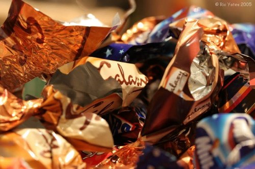 A close-up image of a pile of crushed and crumpled wrappers and packaging materials, primarily made of shiny metallic foil and glossy plastic in various colors including copper, silver, blue, and orange. The wrappers are irregularly shaped, with some displaying partially visible text and branding, and are densely packed together. The background is blurred, suggesting the focus is on the discarded rubbish, which appears to be collected for rubbish removal services possibly in the Twickenham area. The surface underneath the pile is not visible, emphasizing the chaotic arrangement of packaging waste, indicative of typical post-consumer rubbish collected for clearance by Twickenham House Clearance and related waste management companies in the local postcode of Twickenham.
