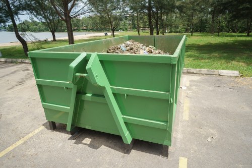 A large, rectangular, green metal skip filled with various types of waste, including paper, cardboard, and mixed debris, situated in an outdoor parking area with yellow line markings. The skip is positioned on a paved surface adjacent to a grassy area with trees and a body of water visible in the background. The skip is supported by black wheels underneath and has a front corner edge that appears slightly tilted, indicating it may not be fully level. The environment suggests a typical waste collection site, with natural light illuminating the scene. The image is relevant to rubbish removal services offered by Twickenham House Clearance in the local area.
