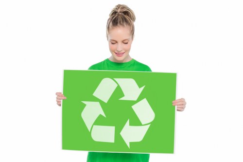 A young woman with blonde hair tied back in a ponytail, wearing a bright green long-sleeved top, holds a large rectangular green sign featuring a white recycling symbol composed of three arrows forming a continuous loop. She is standing against a plain white background, smiling gently, and appears to be promoting environmentally conscious waste disposal. The focus is on the clear, bold recycling graphic and her positive expression, emphasizing the importance of waste sorting and environmentally friendly rubbish removal services in areas such as Twickenham, South West London, provided by Twickenham House Clearance.