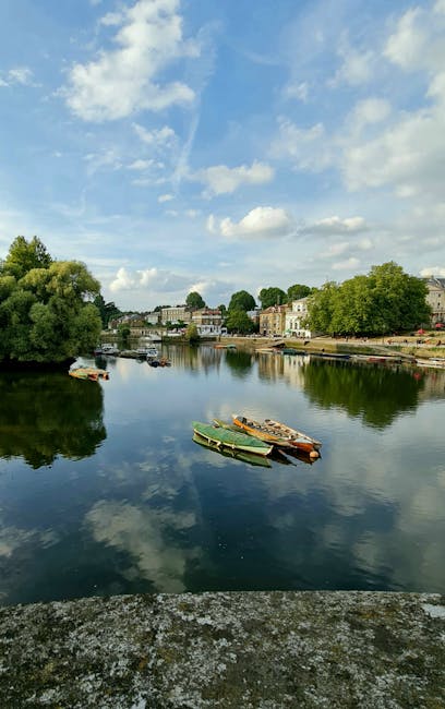 A calm river scene during daytime, with four small boats docked closely together on the reflective water surface, which mirrors the partly cloudy sky and surrounding greenery. The boats, positioned ne