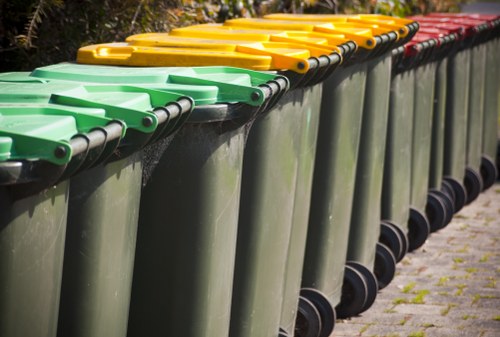 A row of large, wheeled waste bins made of dark green plastic, lined up along the edge of a paved outdoor area. The bins feature hinged lids in bright yellow, indicating different waste separation categories, with the yellow lids at the front and a few red-lidded bins further along the row, all positioned on a cobblestone surface. The background includes some greenery, with trees and bushes, suggesting a residential or community setting typical of Twickenham. The bins are clean, with smooth surfaces, and are systematically arranged, reflecting a professional rubbish collection service such as Twickenham House Clearance operating in the local area for household waste disposal.