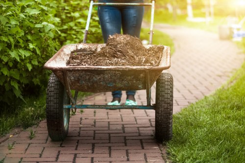 A person wearing blue jeans and dark shoes is pushing a rusted metal wheelbarrow along a paved pathway bordered by green hedges and grass. The wheelbarrow contains dark brown soil or compost, with some loose material spilling over the edges. The pathway appears to be part of an outdoor garden or residential area, with blurred background elements suggesting a sunny day. The scene is indicative of gardening or waste clearance activities, and the image relates to rubbish removal services supported by Twickenham House Clearance in the local area nearby Twickenham.